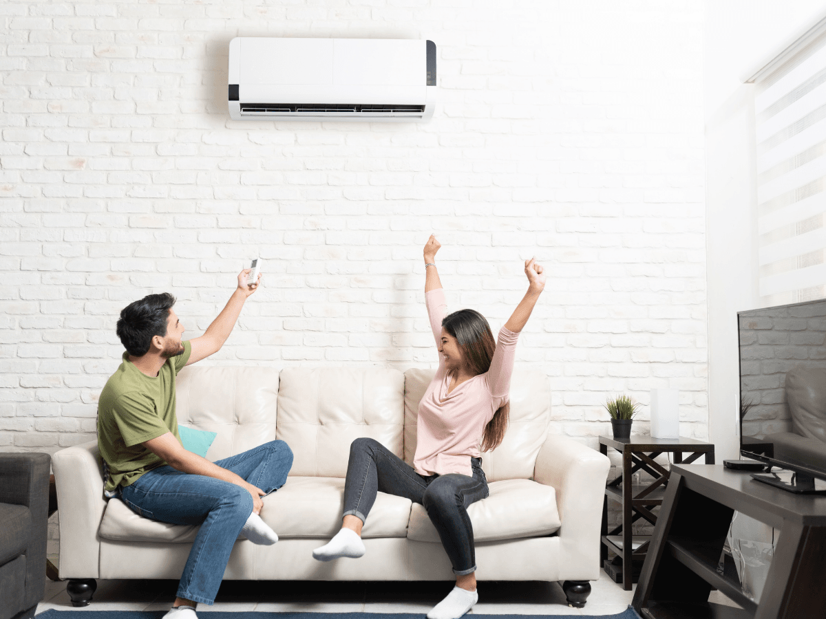 A man and woman sit on a couch in a living room. The man aims a remote at a wall-mounted air conditioner, possibly after a recent ductless mini-split repair, while the woman raises her arms in excitement.