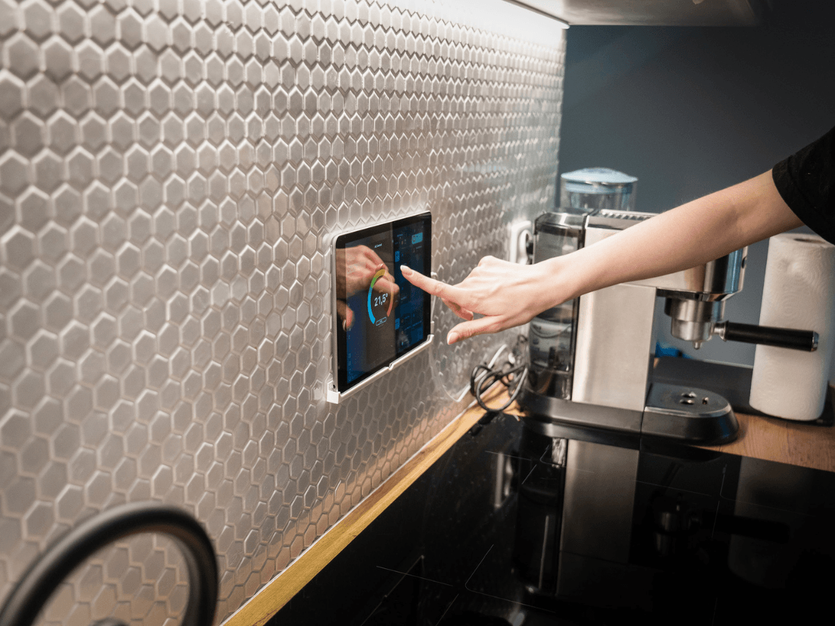 A person uses a wall-mounted touchscreen smart thermostat in a modern kitchen with a hexagonal tile backsplash and coffee machine visible on the counter.