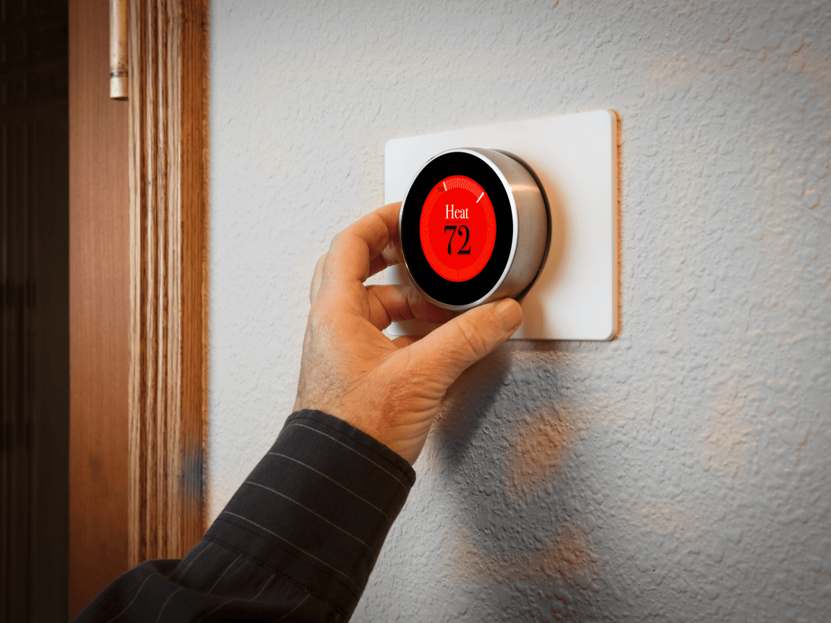 A person adjusts a smart thermostat on a wall, displaying “Heat” and set to 72 degrees.