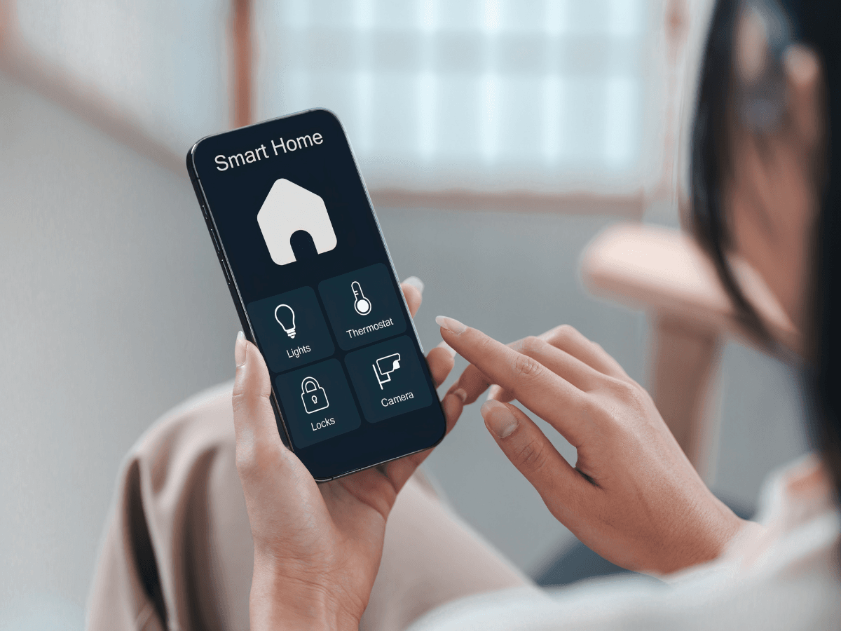 Person holding a smartphone displaying a smart home control app with intuitive options for lights, smart thermostat settings, locks, and cameras.