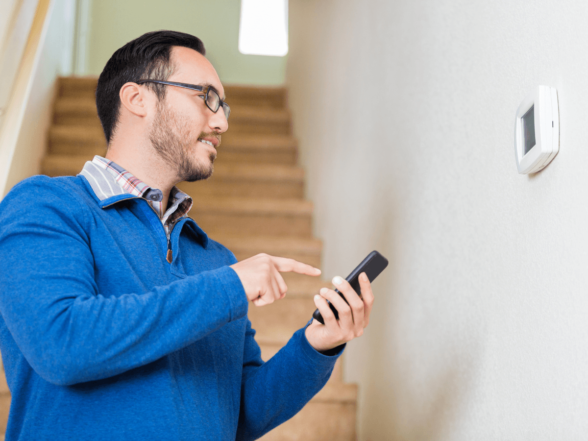 Man standing by stairs adjusts a wall-mounted smart thermostat using a smartphone.