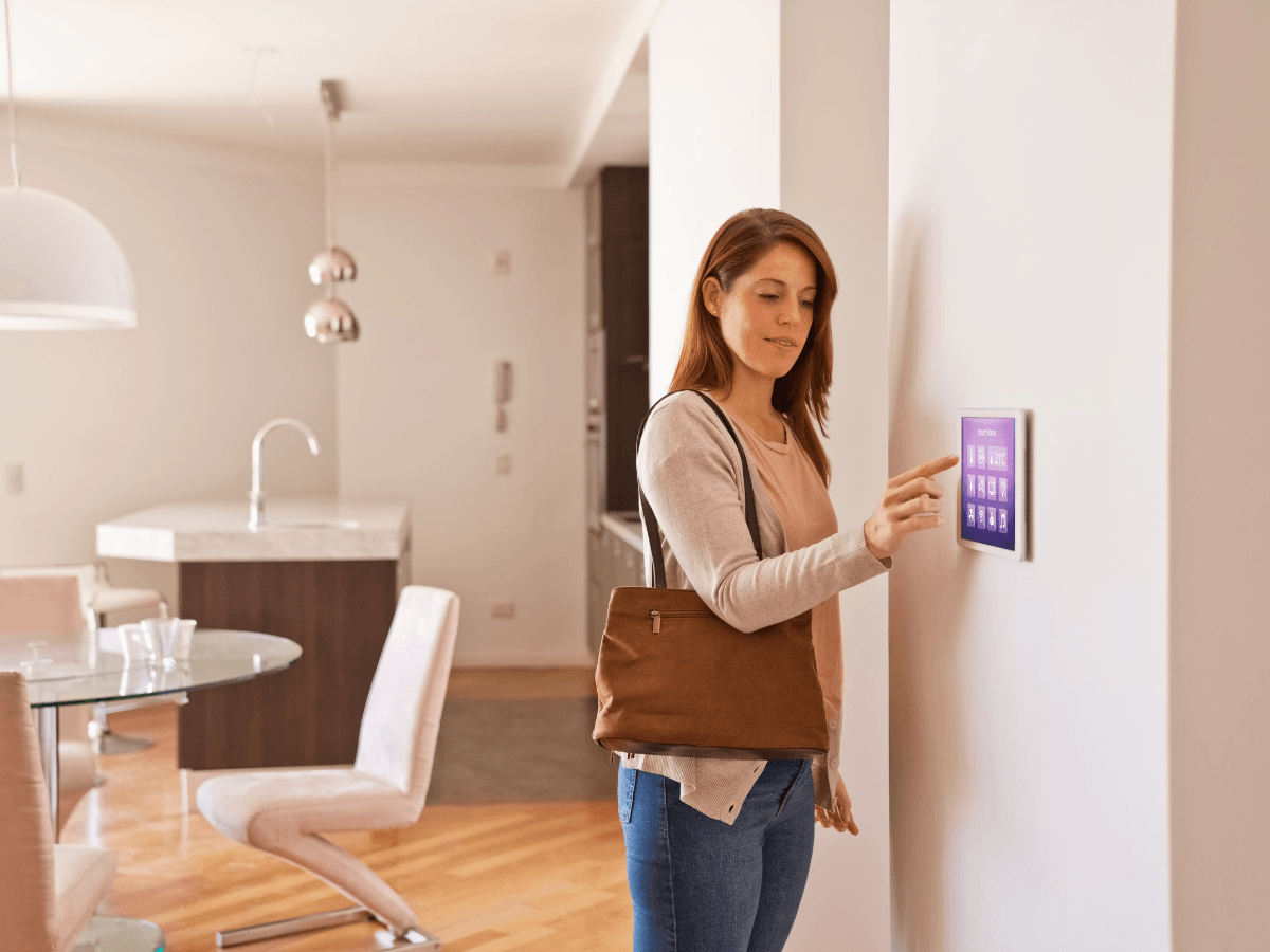 Woman standing in a modern living room, touching a digital home control panel on the wall to adjust the smart thermostat, with a brown shoulder bag and casual attire.