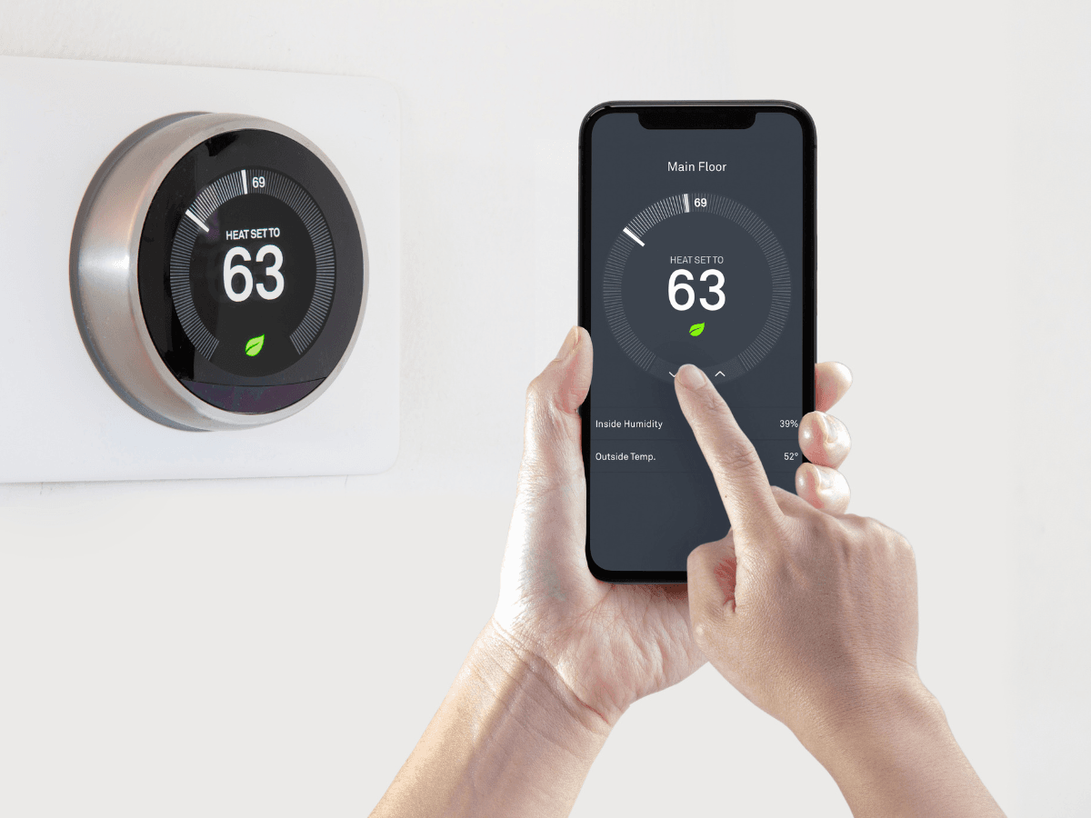 A person uses a smartphone to adjust a smart thermostat on the wall, with both devices displaying a heat setting of 63 degrees.