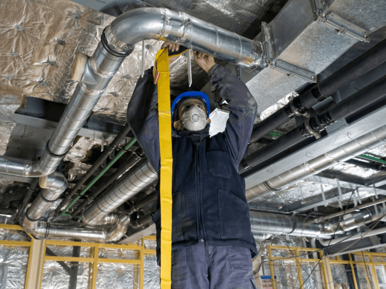 A worker wearing a mask and helmet adjusts metal ductwork on a ceiling in an unfinished building, demonstrating the precision required for ductwork repair Atlanta projects. Rolls of insulation and various pipes are visible around the work area.