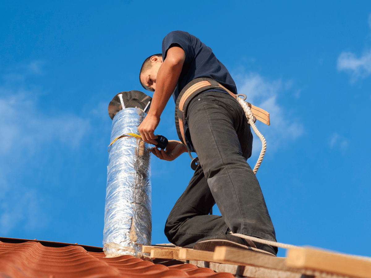A person wearing safety gear secures or installs a metal chimney pipe on a red-tiled roof under a clear blue sky, exemplifying the meticulous attention to detail found in professional ductwork repair Atlanta services.
