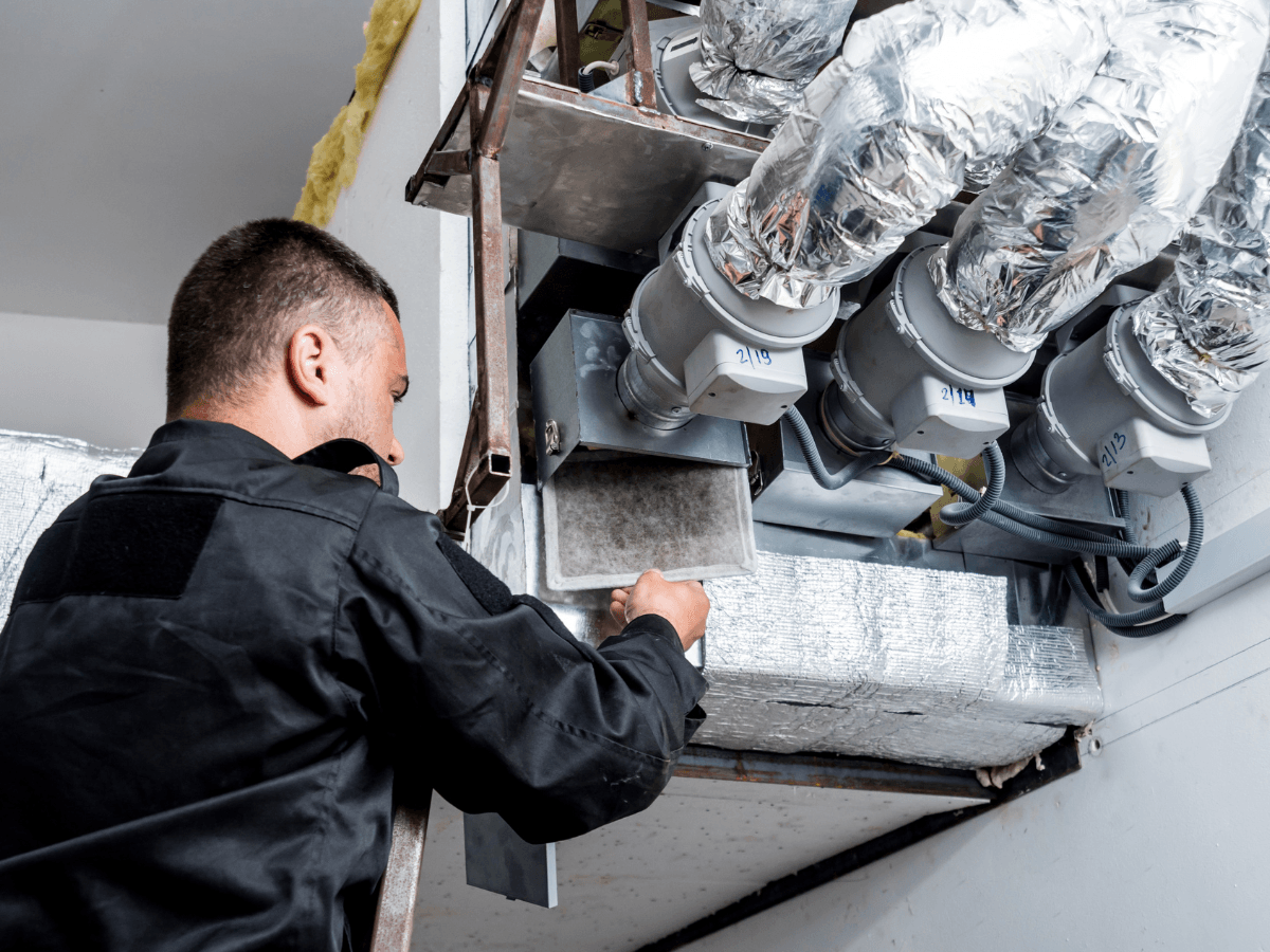 A technician in black workwear checks or replaces a filter in an HVAC air duct system with insulated pipes and electrical components, expertly handling ductwork repair Atlanta residents can rely on.